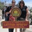 Two women, enjoying a perfect trip, stand beside a sign marking the equator at latitude 00°0000 in Ecuador’s Middle of the World tourist site on a sunny day—making memories that Mom will cherish.
