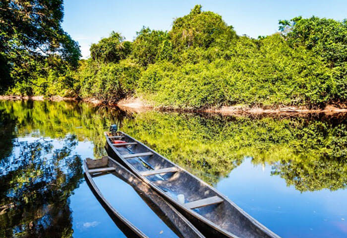 A narrow wooden canoe floats on a calm river surrounded by lush green trees, with clear blue sky and vibrant foliage reflected on the water’s surface—a serene scene among top Venezuela attractions.