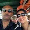 A man and a woman wearing sunglasses smile for a selfie while sitting together on a boat during their South America trip. Bright orange life jackets are visible above them, and other passengers can be seen in the background.