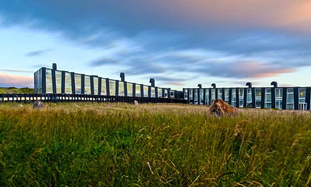 A modern, glass-fronted building—reminiscent of Remota Patagonia—stretches horizontally across a grassy field under a cloudy sunset sky, with warm light reflecting in the windows.
