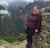 A person stands smiling next to a stone wall on a mountain path in Peru, with lush green mountains and misty clouds in the background—capturing the spirit of travel and adventure.