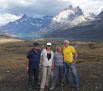 Four people stand together outdoors, smiling, with a scenic background of mountains, a lake, and cloudy skies. Their casual outdoor clothing reflects a stress free adventure, well planned and beautifully executed.