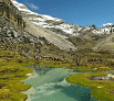 A turquoise river flows through a lush green valley, winding gracefully like a smooth transaction between rocky hills and snow-capped mountains under a clear blue sky.
