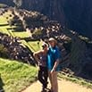 Two people stand smiling on a grassy ledge, enjoying an amazing trip with the ancient ruins of Machu Picchu visible in the sunlit valley below, surrounded by lush green mountains.