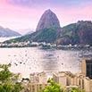 A scenic view of Rio de Janeiro’s Sugarloaf Mountain at sunset, with well-organized boats on the water and city buildings in the foreground. The sky is tinged with pink and purple hues.