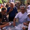 Four older men sit around a table outdoors playing cards, while another man stands nearby watching—perhaps discussing travel planning or organising our trip. People and trees in the background suggest a park or public gathering.