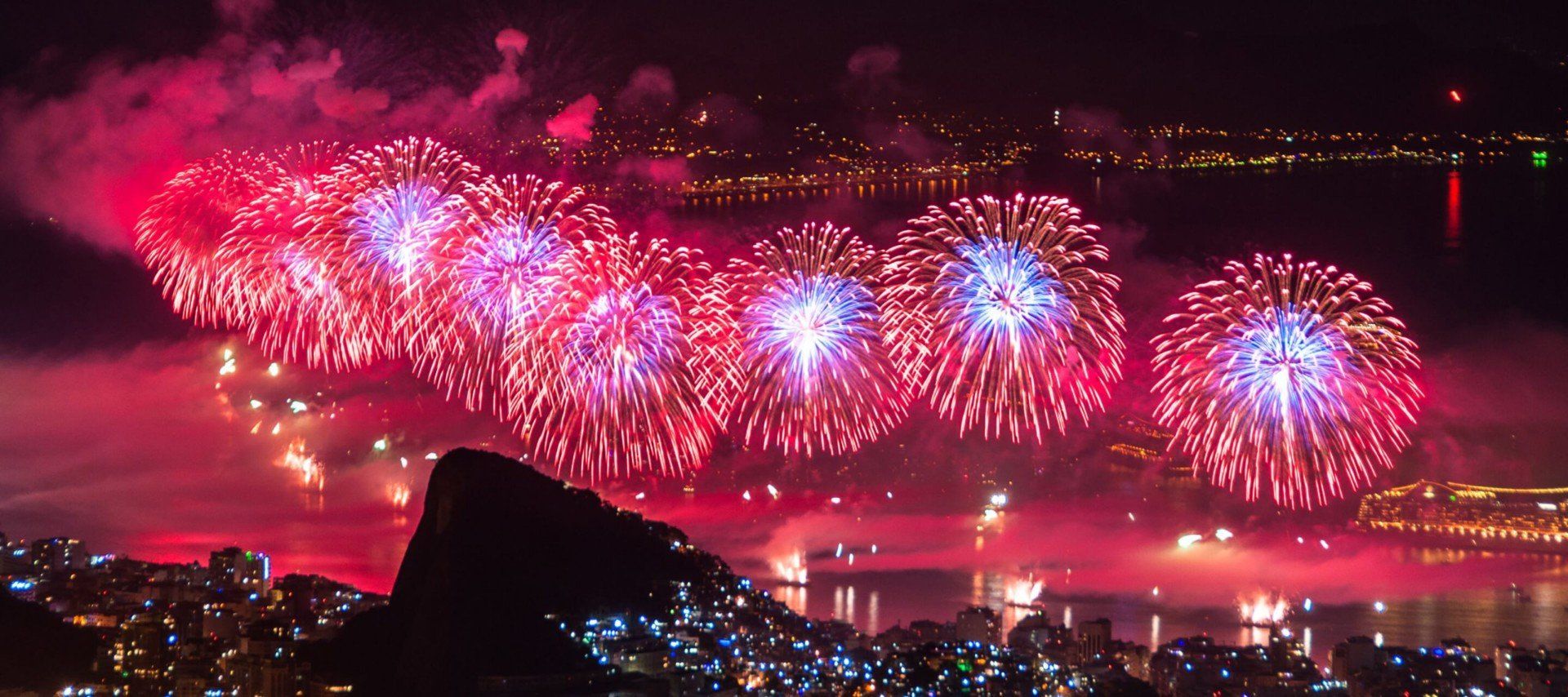 Bright fireworks in red and blue explode over a city waterfront at night, illuminating the water and city lights below; mountains and reflections are visible in the foreground, capturing the magic of Rio New Years Eve celebrations.