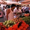 Two people examine produce at an outdoor market stall filled with apples, tomatoes, and other fruits and vegetables; everything went smoothly as shoppers stroll by under striped canopies in the background.