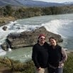 Two people stand close together and smile in front of a river with rapids, surrounded by greenery and mountains—their adventure clearly went above and beyond their expectations.