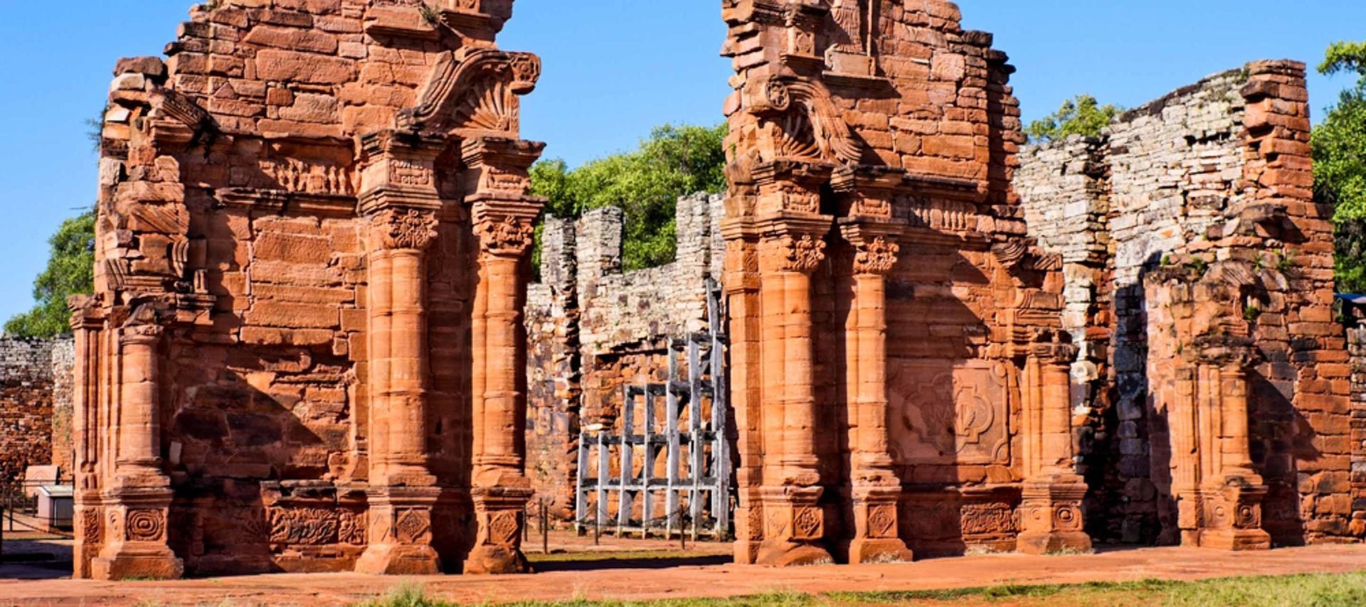 Ancient red stone ruins with tall columns and detailed carvings, partially standing walls, and a metal gate. Set outdoors under a clear blue sky with greenery, San Ignacio Mini is a Jesuit Mission explored on guided tours.