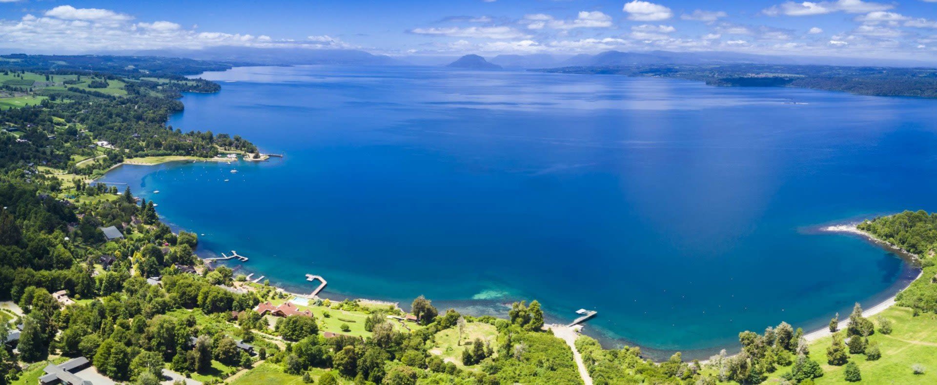 Aerial view of a large, blue lake in Chile surrounded by lush green hills, with small docks and scattered buildings—an inviting scene for visitors exploring beyond just Chile visa requirements under a bright, partly cloudy sky.