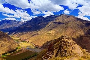 Mountain landscape under a bright blue sky with scattered clouds; green valley and fields below, rugged hills and ancient stone ruins in the foreground—capture the wonder of Amazing Peru on your next Peru travel adventure.