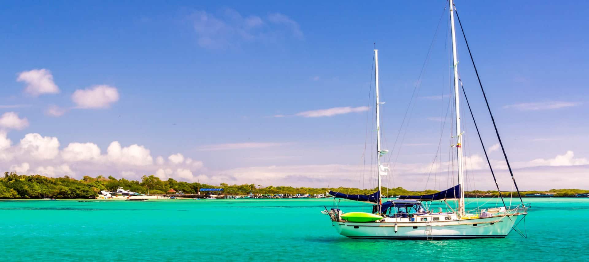 A white sailboat floats on vibrant turquoise water near a green shoreline under a bright blue sky with scattered clouds—the perfect scene for an unforgettable Ecuador honeymoon. Other boats are docked in the distance.