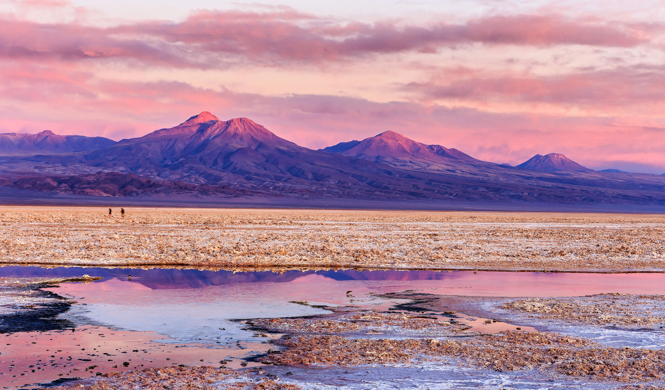 Pink and purple sunset over distant mountains, a wide salt flat in the foreground with shallow reflective water, and two small people walking across the landscape—an unforgettable scene among top Chile attractions.