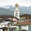 A small town with a prominent church tower featuring a large cross, surrounded by rustic buildings with red-tiled roofs and green hills, creates the perfect backdrop for an exceptional trip under a cloudy sky.