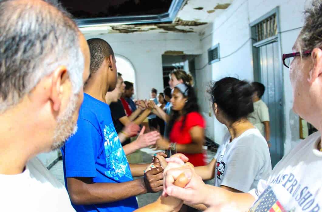 A group of people stand in two lines facing each other, holding hands and clapping, engaging in a lively group activity indoors under a damaged ceiling—a unique scene among the many vibrant things to do in Colombia.