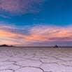 Cracked desert ground stretches into the distance under a colorful sky at sunset, with hues of pink, purple, and blue. Mountains and a solitary small structure are visible on the distant horizon.