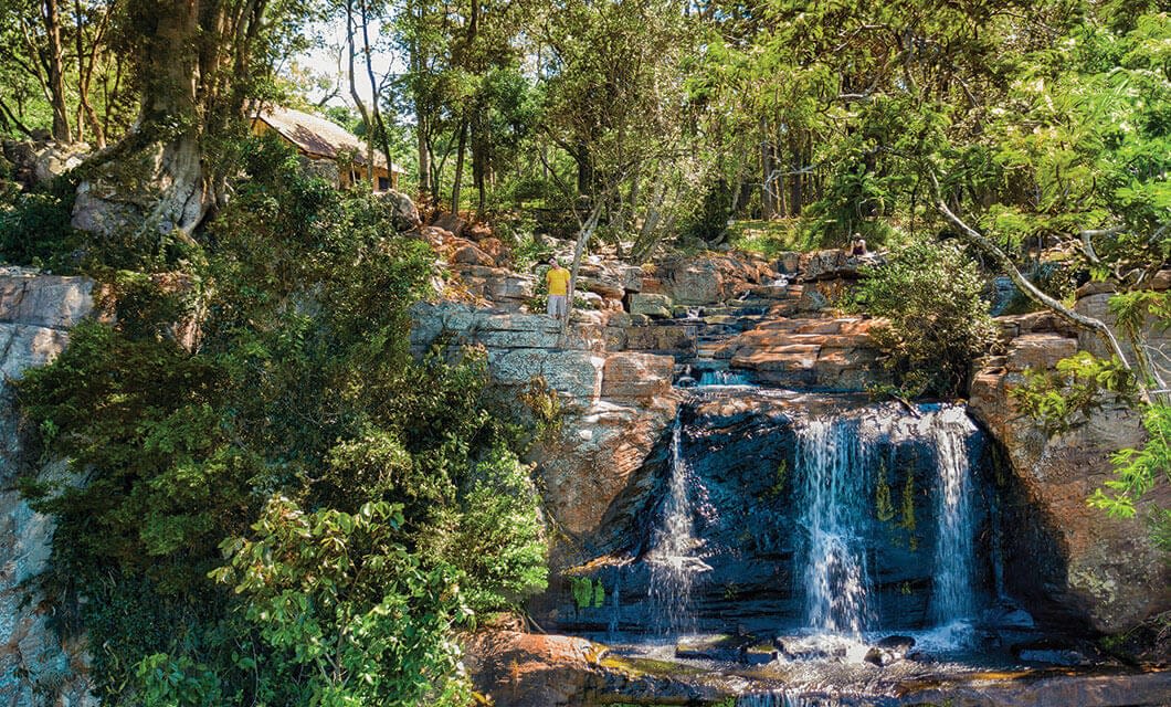 A small waterfall cascades over rocks surrounded by lush green trees and vegetation, with a person in yellow standing on the rocks and a thatched-roof hut partially visible—one of the unique things to do in Paraguay.
