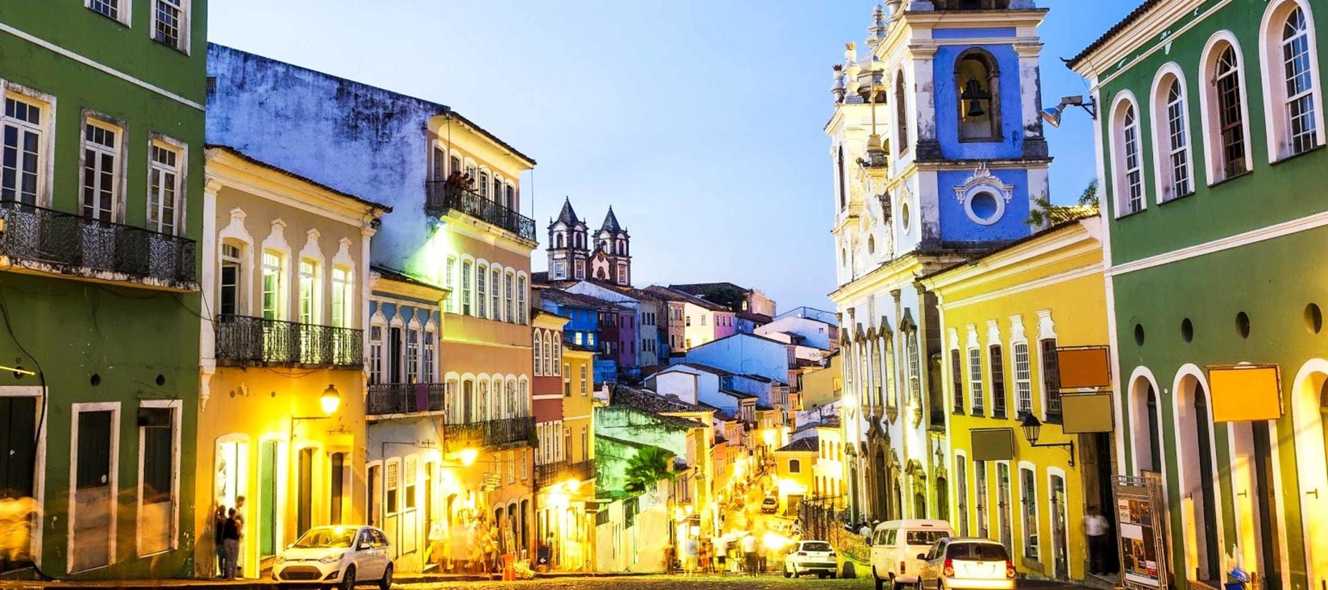 A vibrant street scene in Pelourinho, Salvador da Bahia, at dusk, showcases colorful colonial buildings, glowing streetlights, cars, and people strolling along the cobblestone road—a perfect setting for unforgettable Bahia Tours.