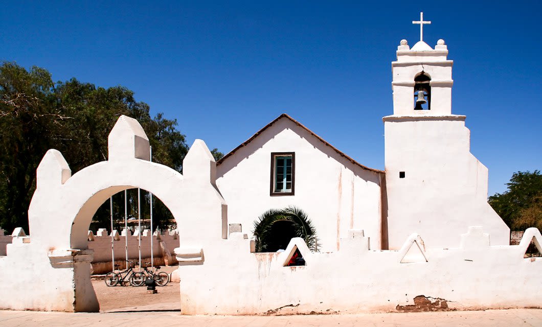 A white adobe church with a bell tower and a cross sits under a clear blue sky in the Atacama Desert. An arched entrance and a low wall with geometric shapes frame the front, with Awasi Atacama’s bicycles resting inside the courtyard.