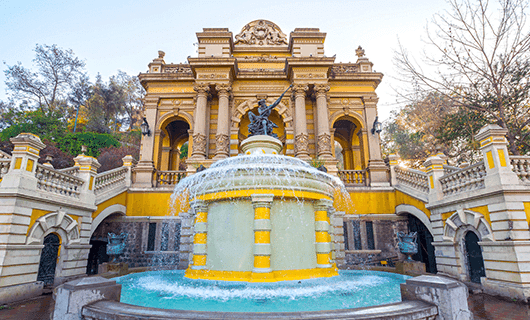 A grand yellow and white neoclassical fountain with statues and water features sits before an ornate building at this luxury hotel in Chile, featuring arches, columns, decorative sculptures, and flanked by trees—reminiscent of Viña Vik Hotel’s elegance.