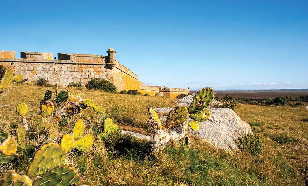 An old stone fortress with a small turret stands on a grassy hill, surrounded by yellow-green cacti. Beneath the clear blue sky, this scenic spot is one of the unique things to do in Uruguay as the landscape stretches into the distance.