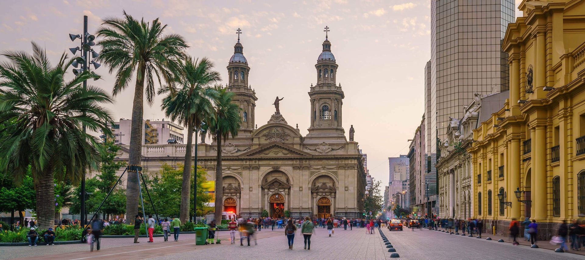 A busy plaza with palm trees and people walking, featuring a grand historic cathedral with twin towers in the background, surrounded by yellow and modern buildings at sunset—a perfect stop for Chile Train Tours.