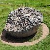A large, oval-shaped stone with intricate carvings and geometric patterns sits smoothly on a grassy area, surrounded by a circular dirt path—inviting visitors to pause during their trip along the metal fence.