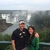 A smiling couple poses together in front of a railing with large waterfalls and lush greenery in the background, clearly having a great time all around.