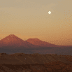 Desert landscape at sunset with two distant mountains, a pale sky, and a bright full moon above the horizon—evoking the magic of a trip across Chile or Argentina, where warm, golden hues bathe the breathtaking scene.