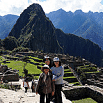 Three people pose together, smiling, at the ancient Inca site of Machu Picchu in Peru's Sacred Valley. Stone terraces and buildings surround them, with a tall mountain rising dramatically in the background under a partly cloudy sky.