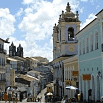Colorful colonial buildings line a cobblestone street under a bright blue sky with fluffy clouds; a historic church with a clock tower stands prominently in the center, where visitors enjoy excellent service and competitive rates nearby.