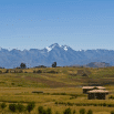 A small house sits in a grassy field with rolling hills in the foreground and snow-capped mountains beyond—a perfect view for booking our trip under a clear blue sky.