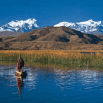 A travel agent watches as a person in traditional clothing paddles a boat on a calm lake, surrounded by tall reeds, with brown hills and snow-capped mountains under a clear blue sky in the background.