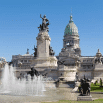 A large ornate building with a green dome and classical columns stands behind a monument with statues and a super fountain in the foreground under a clear blue sky.
