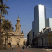 A city square with people walking, featuring a historic church with twin bell towers on the left, palm trees, and a modern glass skyscraper in the background—perfect for a once in a lifetime trip under a clear blue sky.