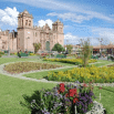 A sensational colonial-style church stands behind a well-maintained garden, bursting with colorful flowers and winding green paths under a bright blue sky with scattered clouds.