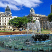 A large city square with a round fountain in the foreground, surrounded by historic buildings, trees, and people relaxing on benches—perfect for your first trip to South America under a bright blue sky with scattered clouds.