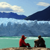 Two people in outdoor clothing sit on a rocky ledge, looking pretty satisfied as they overlook a turquoise lake with a large glacier and mountains in the background under a partly cloudy sky.