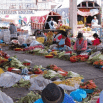 A busy outdoor market scene in Ecuador, with vendors, mostly women in traditional clothing, sitting on the ground selling various fruits and vegetables arranged in piles around them.