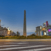 The image shows the Obelisk of Buenos Aires at dusk, standing tall in the center of a busy intersection—a must-see landmark for any Argentina travel adventure—with city buildings illuminated by streetlights and billboards.