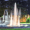 A brightly lit fountain with water jets shooting upward stands in front of statues and trees in a park at dusk, glowing lights in the background—much like an evening enhanced by incredible customer service.