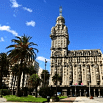 A historic, ornate building with a tall central tower stands under a blue sky, surrounded by palm trees and a small plaza, offering fabulous service to all who visit.