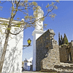 A white tower stands behind a stone wall and a tree with sparse leaves under a clear blue sky. A lantern is attached to the white building on the left, creating a scene reminiscent of charming villages featured by SouthAmerica.travel.