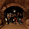 A group of people stands together inside a dimly lit brick wine cellar, surrounded by large wooden barrels on either side, as part of an organized tour.