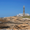 A tall lighthouse with horizontal stripes stands on rocky terrain near the ocean, beside a white building, under a clear blue sky—an inspiring sight for your South America travel suggestions.