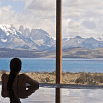 A person in a pool has a fabulous time gazing out a large window at blue water, rugged terrain, and snow-capped mountains beneath a partly cloudy sky.