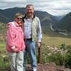 A man and woman stand together outdoors on a grassy hill in Peru with mountains and a winding river behind them, both wearing casual clothes and jackets, smiling at the camera during their well-planned trip organization.