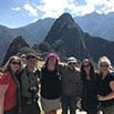 A group of six people pose together, smiling, in front of the ancient ruins and jagged peaks of Machu Picchu on a bright, sunny day during their amazing trip.