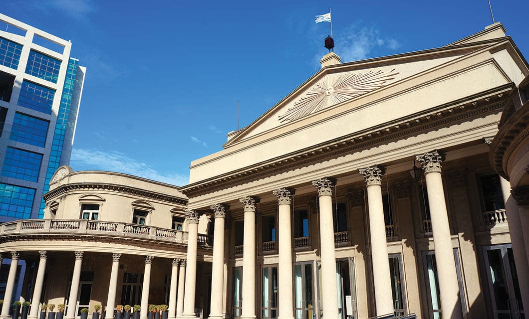 A neoclassical building with tall columns and a triangular pediment, topped by a flag—a striking example of Uruguay attractions. To the left, a modern glass building and a rounded section of the older structure rise under a clear blue sky.
